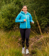 Woman running outdoors in a blue jacket and black leggings with greenery in the background